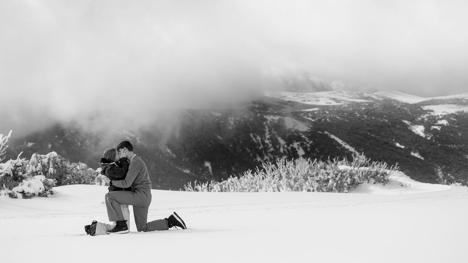 A couple share a kiss in the snow, with snow  covered mountains in the distance with mountains and trees in stunning winter landscape.
