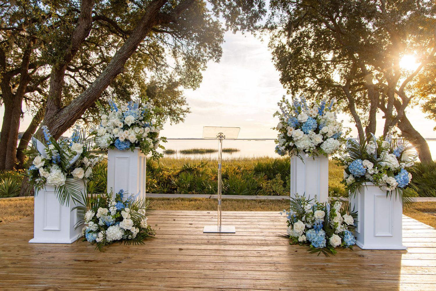 Outdoor wedding ceremony setup with white chairs and floral arrangements under oak trees at sunset.