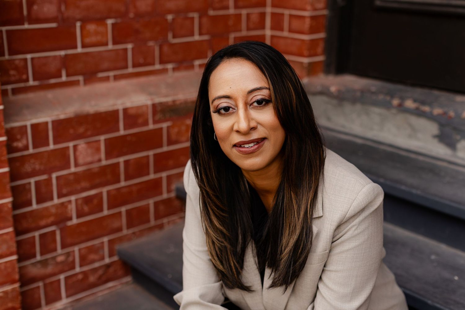 Professional portrait of someone in a cream blazer sitting on steps against a red brick wall background.