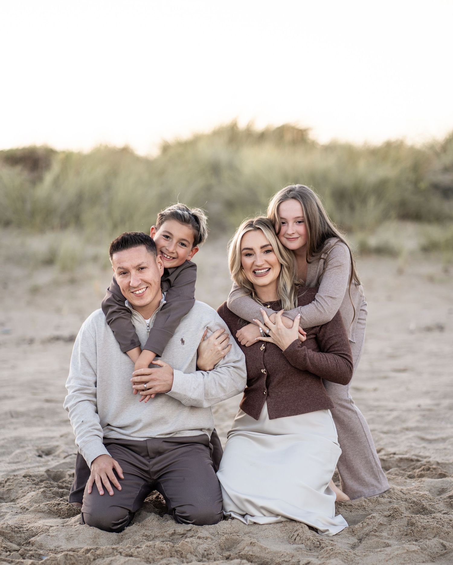 A couple in casual attire shares tender moments while sitting together on a sandy beach at sunset.