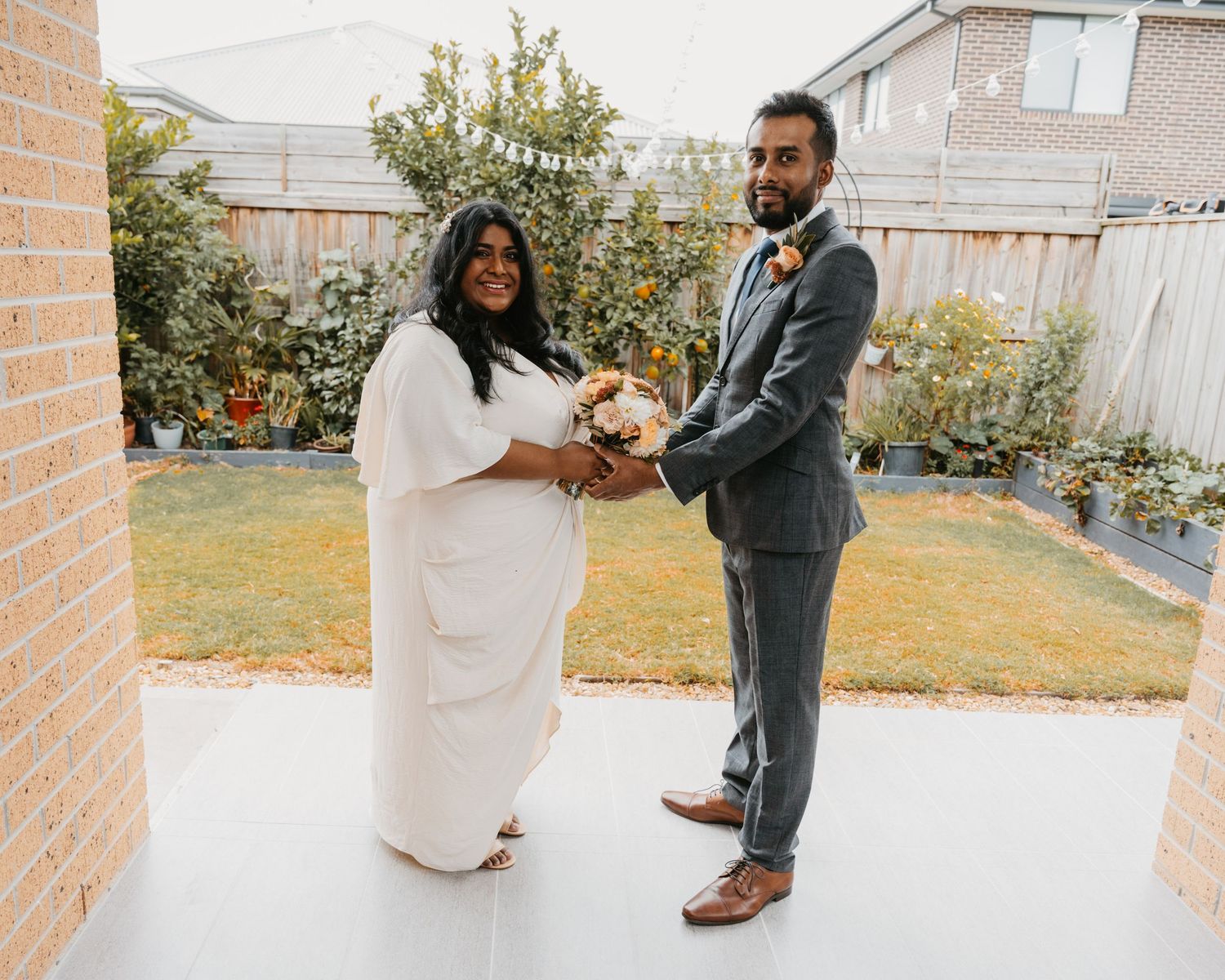 A couple in formal attire dances together in a backyard garden setting during a wedding celebration.