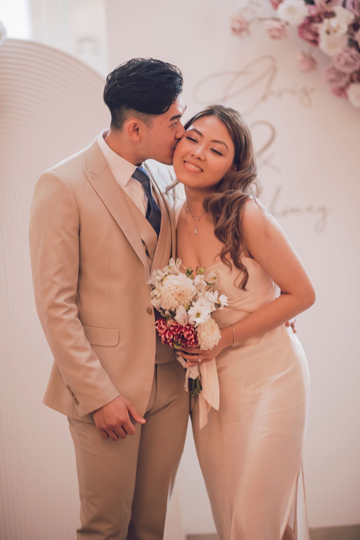 Romantic couple in beige formal attire share intimate moments in a soft pink-toned photo series.