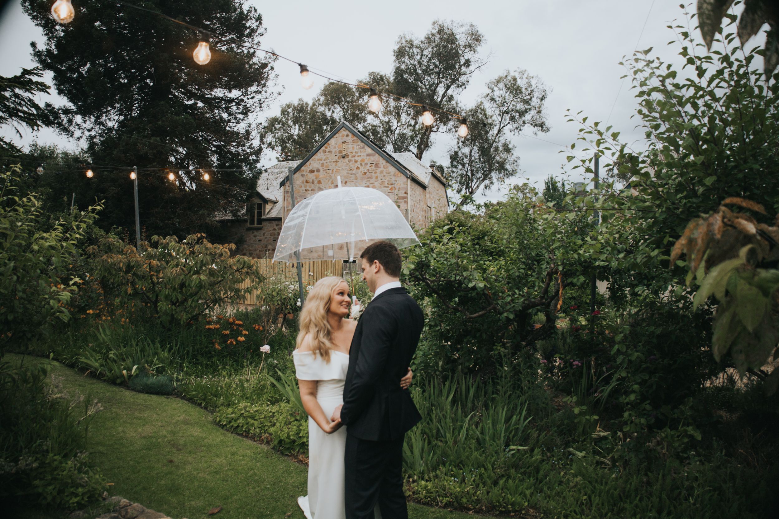 Bride and groom sharing a romantic moment under a clear umbrella in a lush garden.