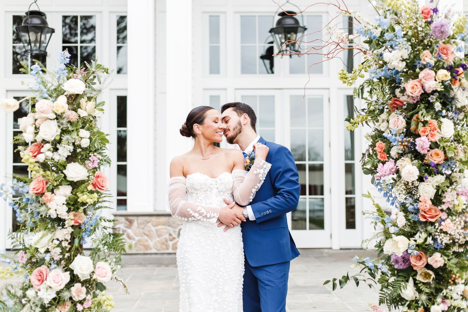 A wedding couple poses under an elegant floral arch with pink and white roses against a white building exterior.