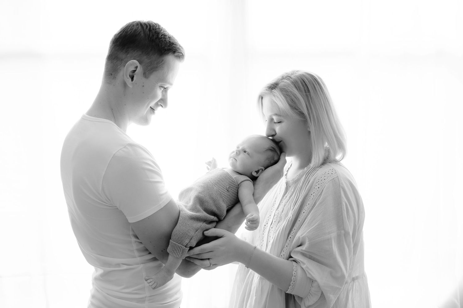 Maminka dává pusinku na hlavičku newborn narozeného miminka, které drží v náruči tatínek a všichni jsou spolu doma.