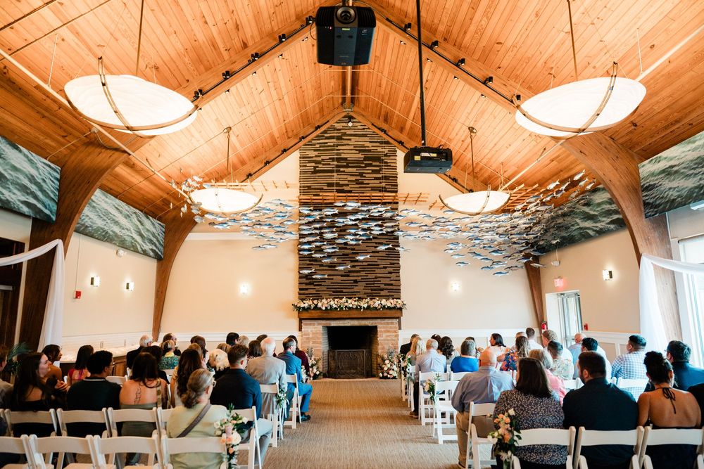 Modern rustic wedding venue interior with exposed wooden beams, white walls and pendant lighting fills with guests seated.