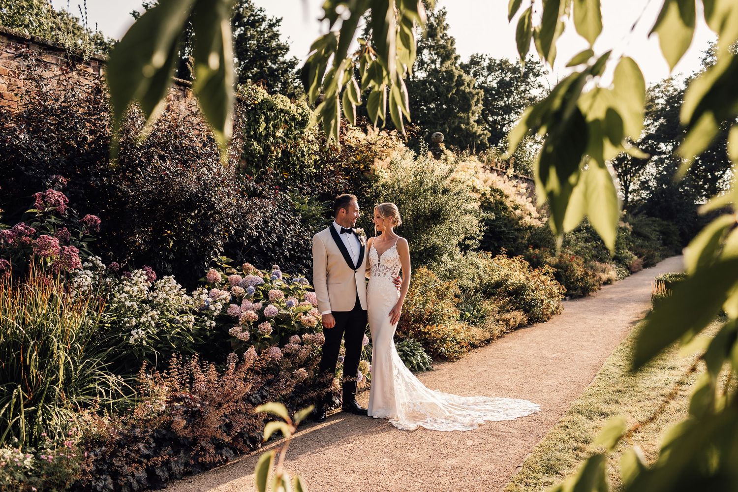 A couple in wedding attire stands on a garden path surrounded by lush summer blooms and greenery.