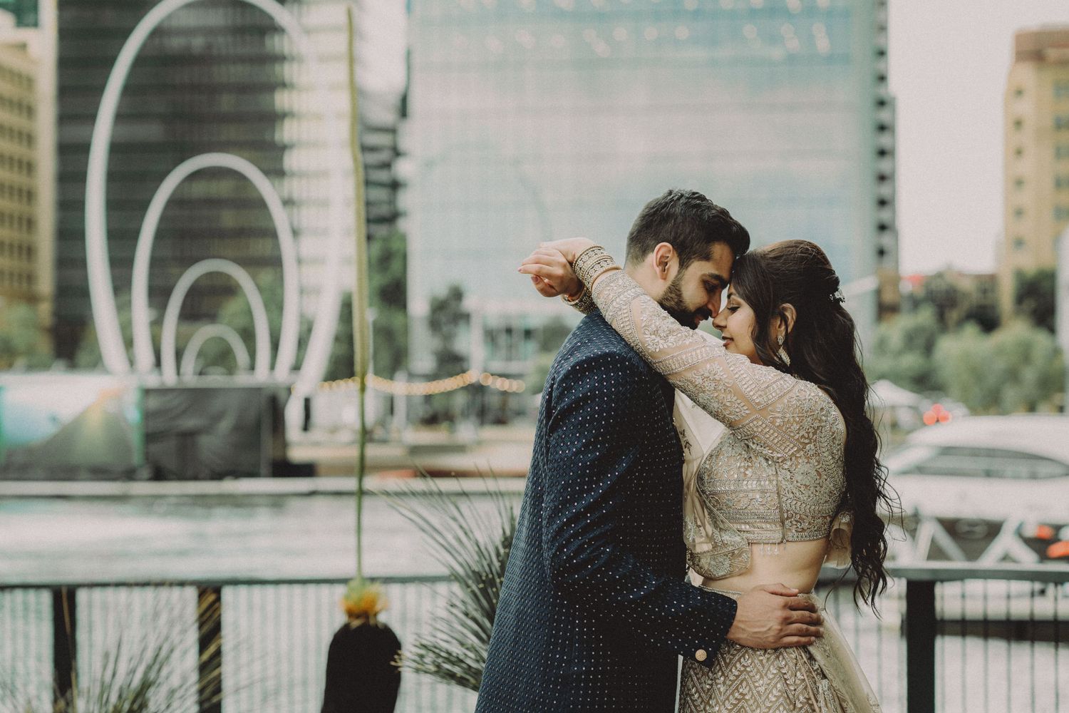 A romantic couple embraces along a waterfront promenade with city buildings in the background.