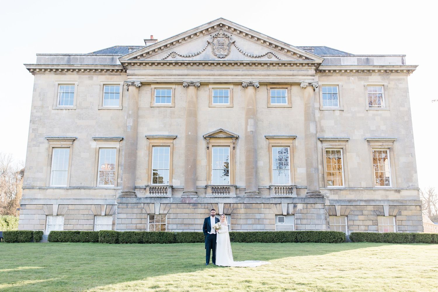 Elegant couple stands together on the lawn of a grand historic mansion with classical architecture in the background at Botleys Mansion