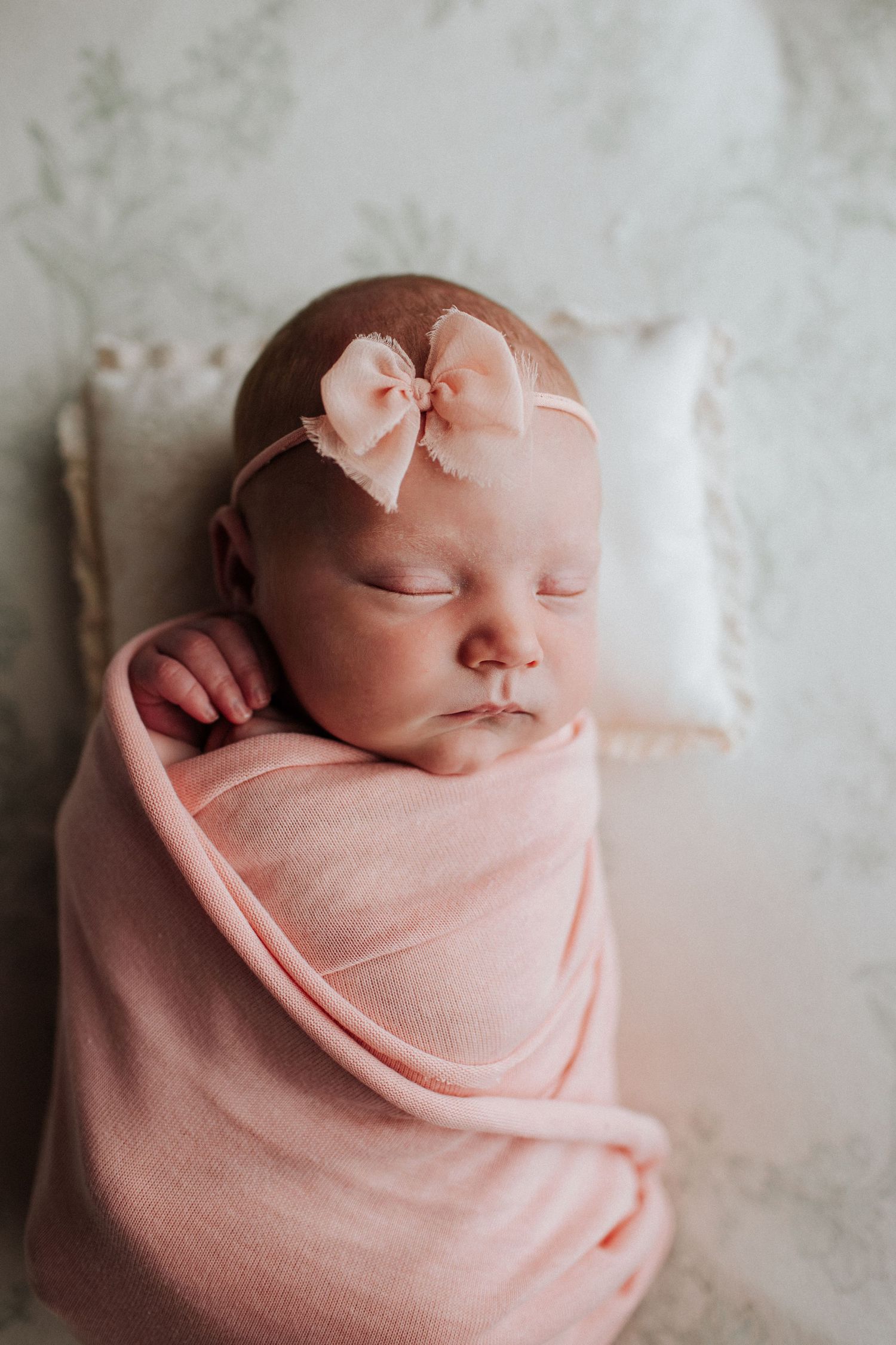 Sleeping newborn baby girl swaddled in pink wrap with bow headband resting on white pillow.