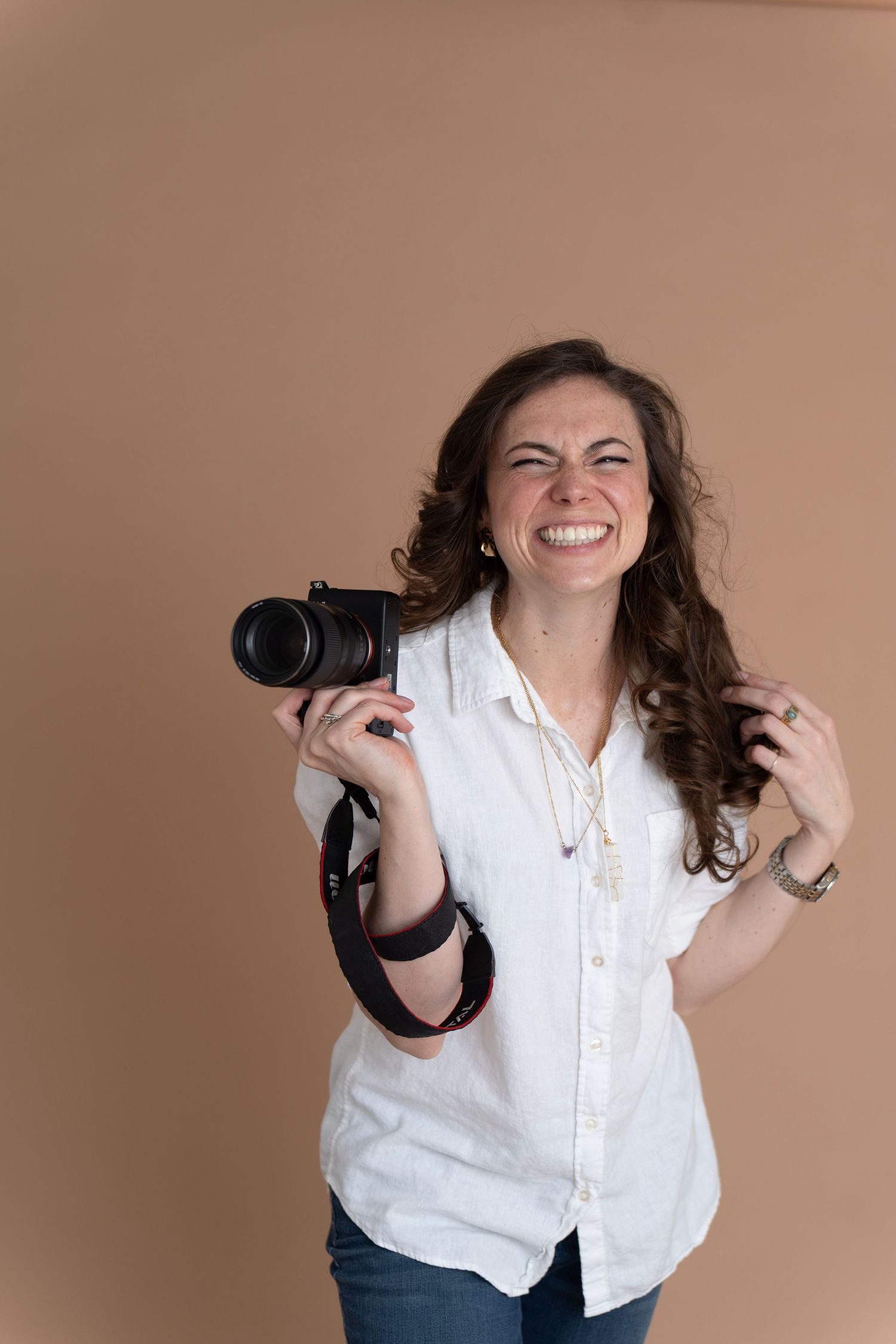 Woman in white shirt holding a DSLR camera on her shoulder, posing confidently against a tan background.