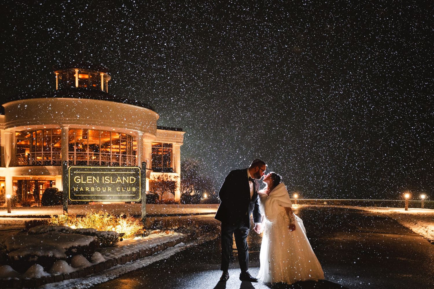 Glen Island Harbour Club wedding venue in New Rochelle during a snowstorm with kissing bride and groom on driveway at night.