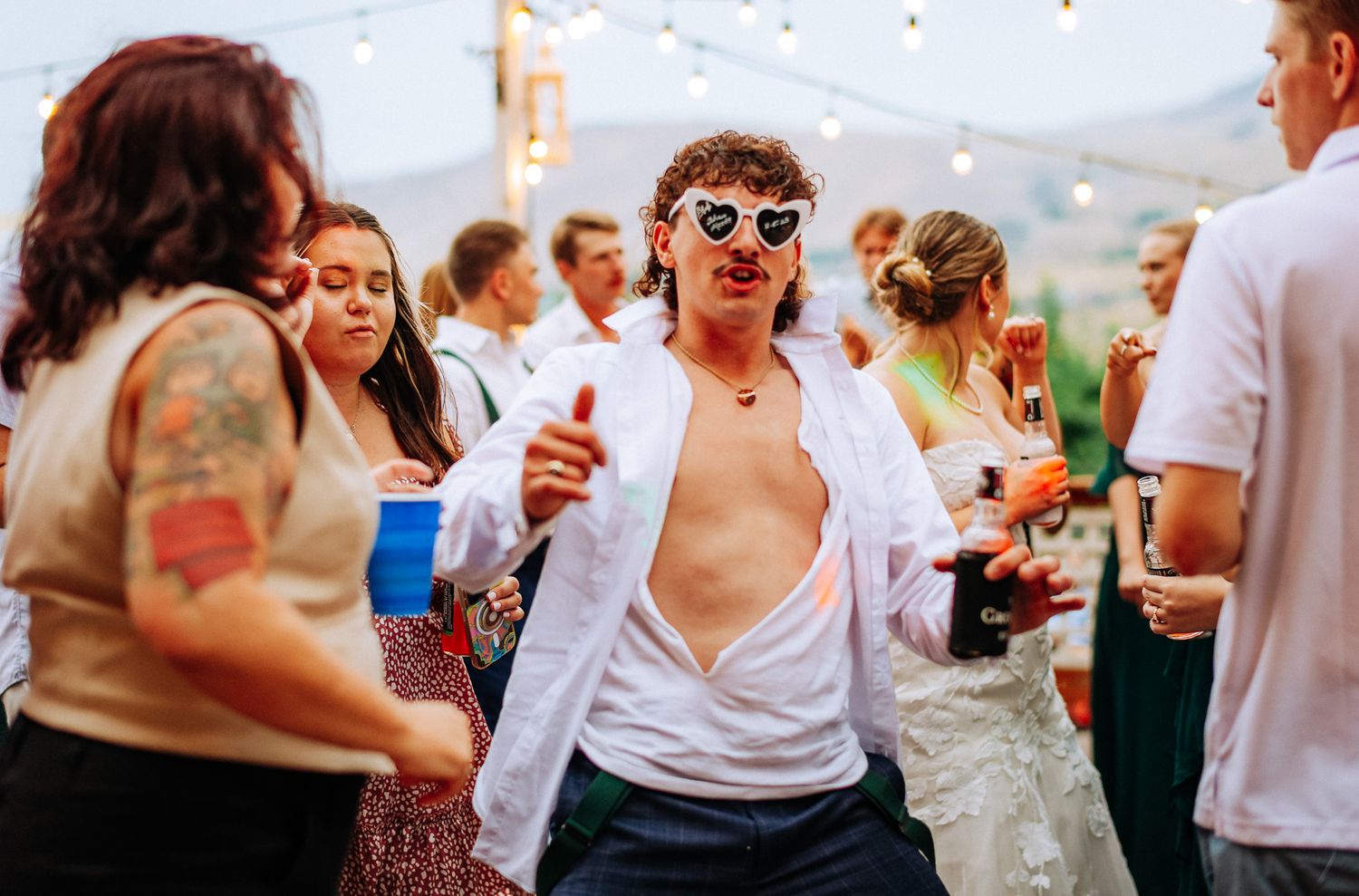 Man in heart sunglasses and open white shirt dancing at an outdoor party with string lights.