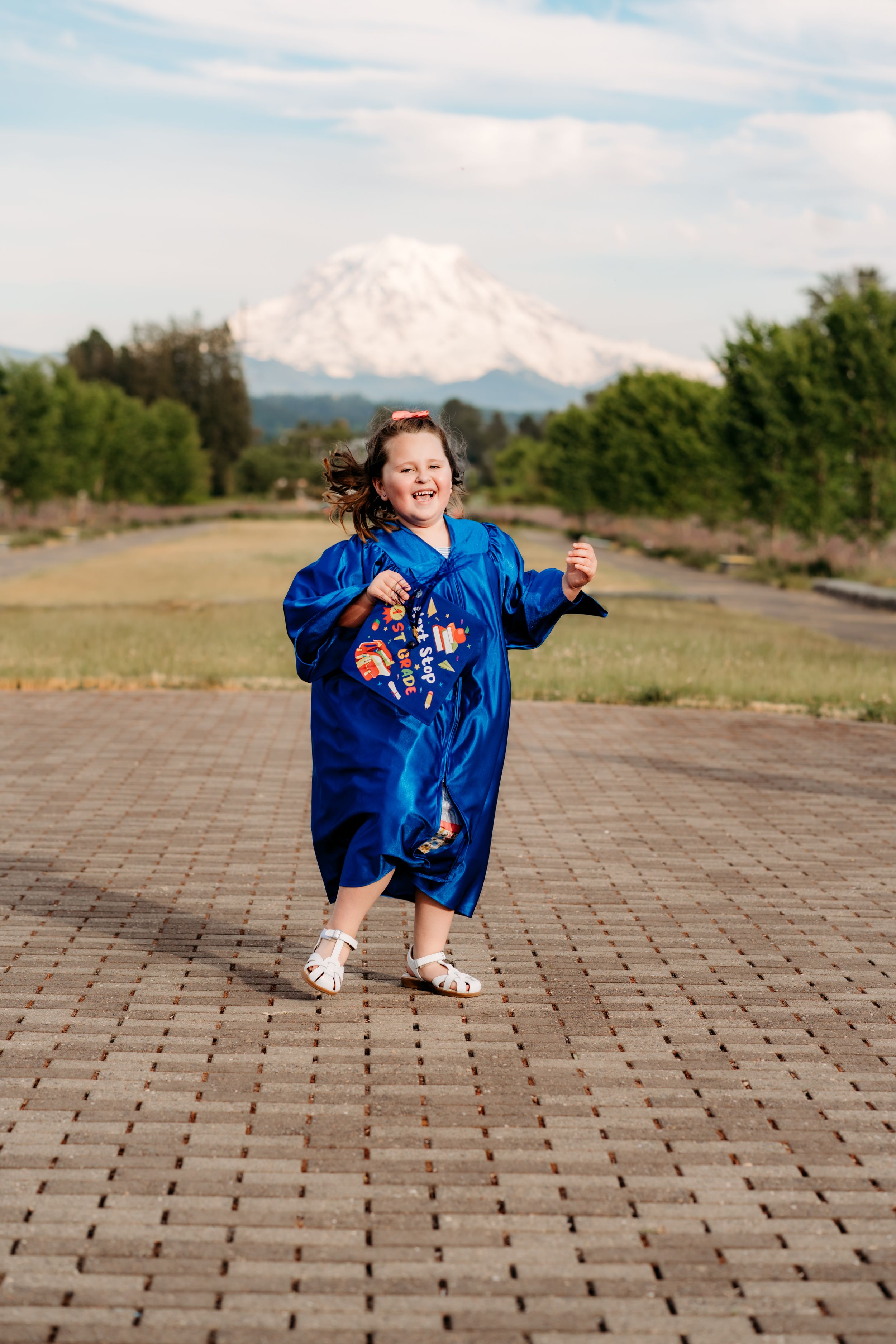 Young girl in blue graduation gown dancing on brick path with snow-capped Mount Rainier in background.
