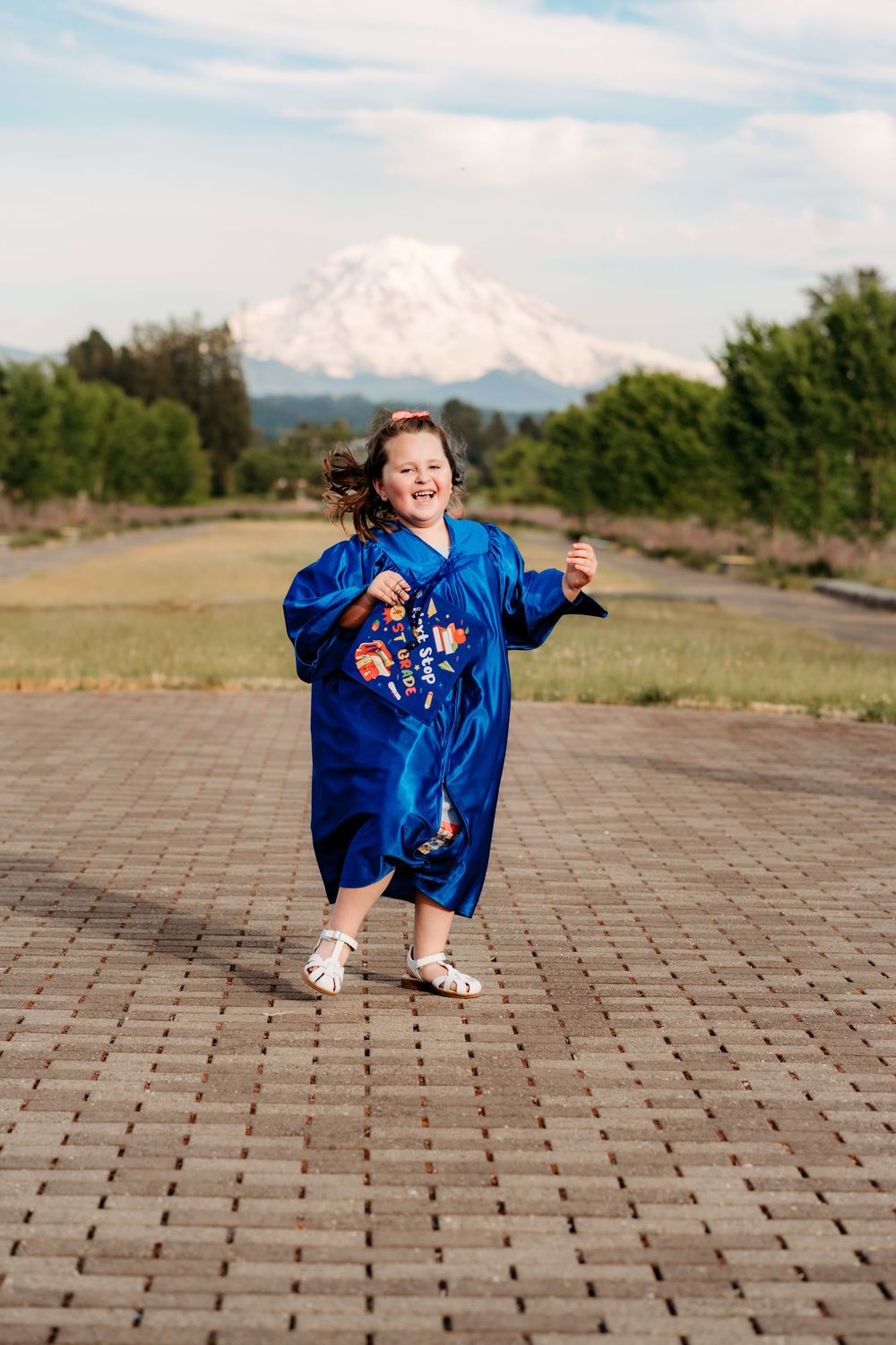Young girl in blue graduation gown dancing on brick path with snow-capped Mount Rainier in background.
