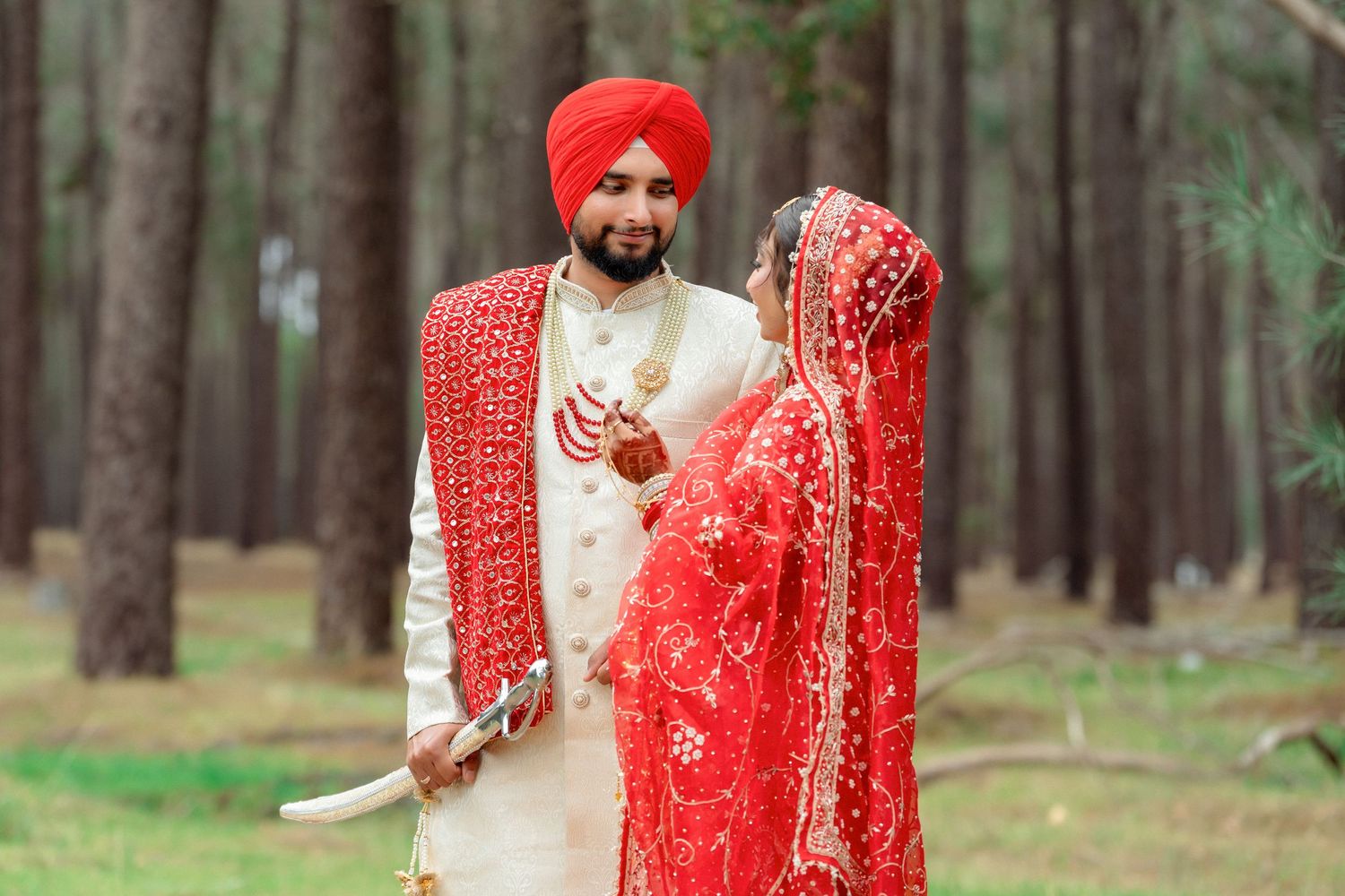 Sikh bride and groom in traditional red and gold wedding attire standing in a pine forest.