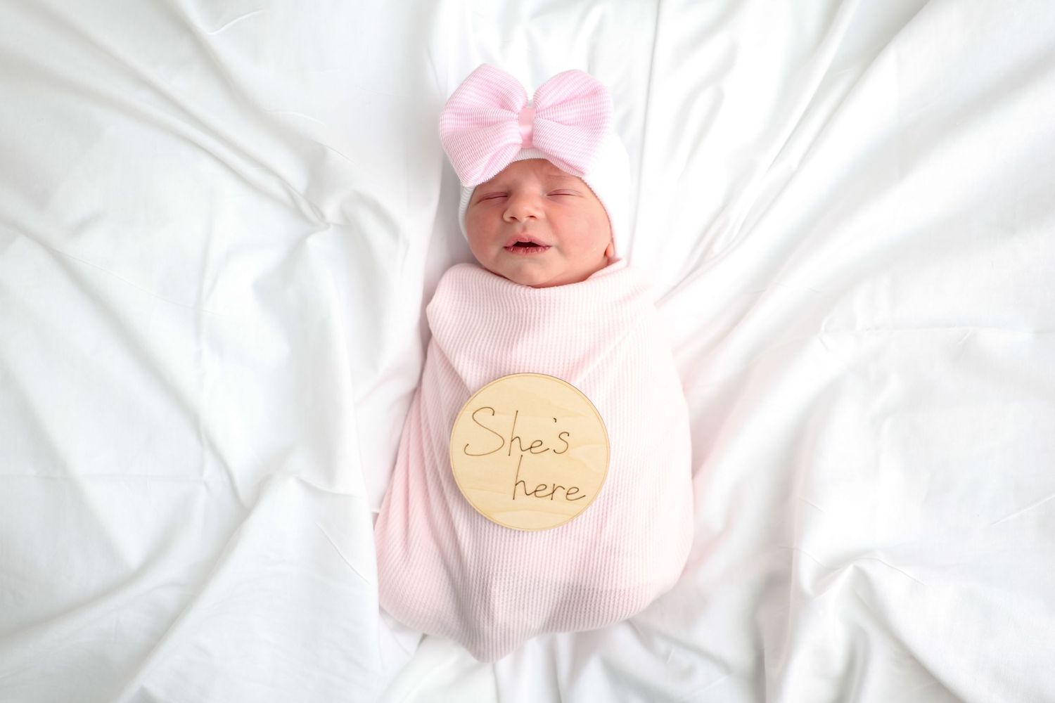 Newborn baby girl swaddled in pink blanket with bow headband and She's Here milestone disc.