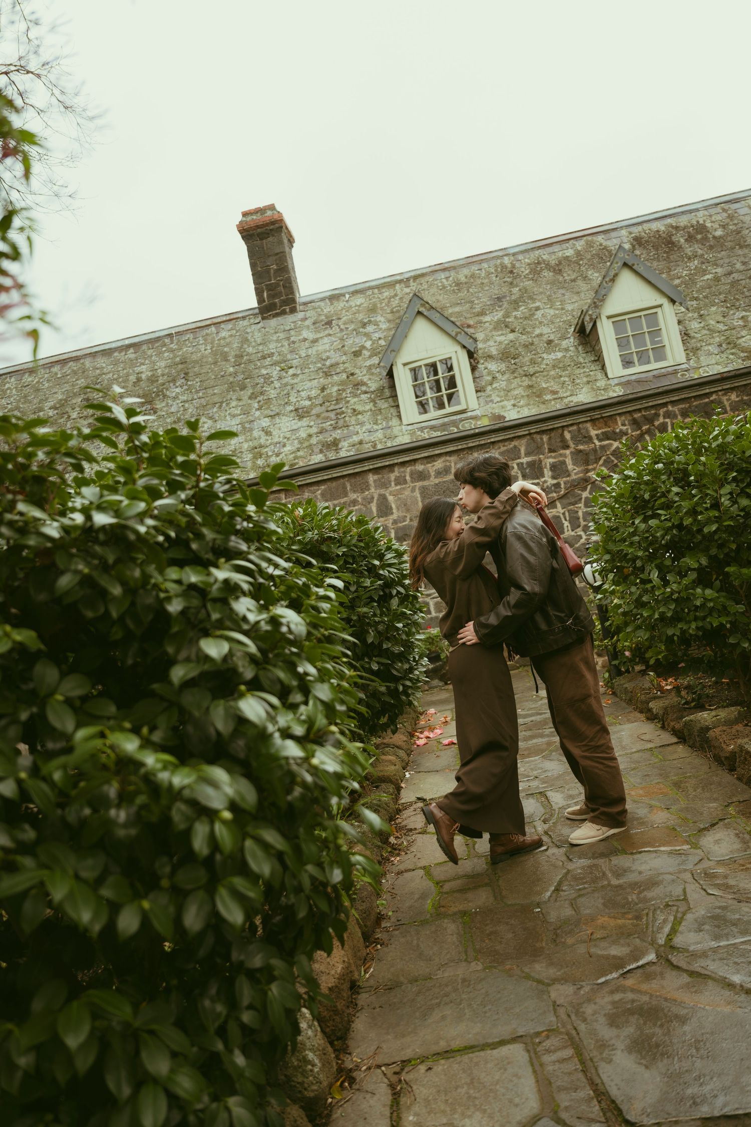 Couple embracing on stone path beside lush hedges outside historic stone building with dormer windows on overcast day.