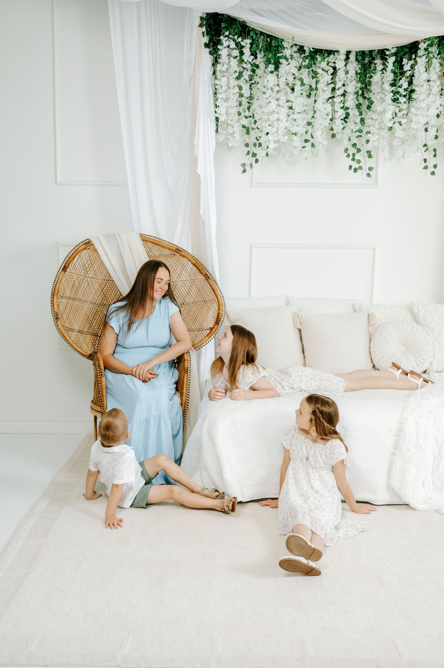 A pregnant woman in a blue dress sits in a wicker chair surrounded by three children in a bright white room.