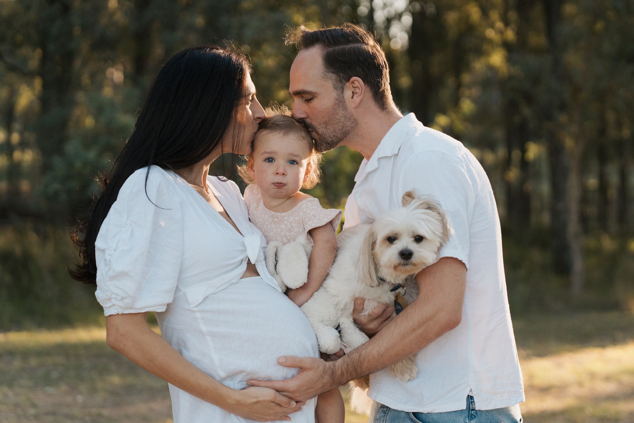 Pregnant woman in white dress with husband holding toddler and white dog outdoors in golden hour light Rouse hill family photoshoot