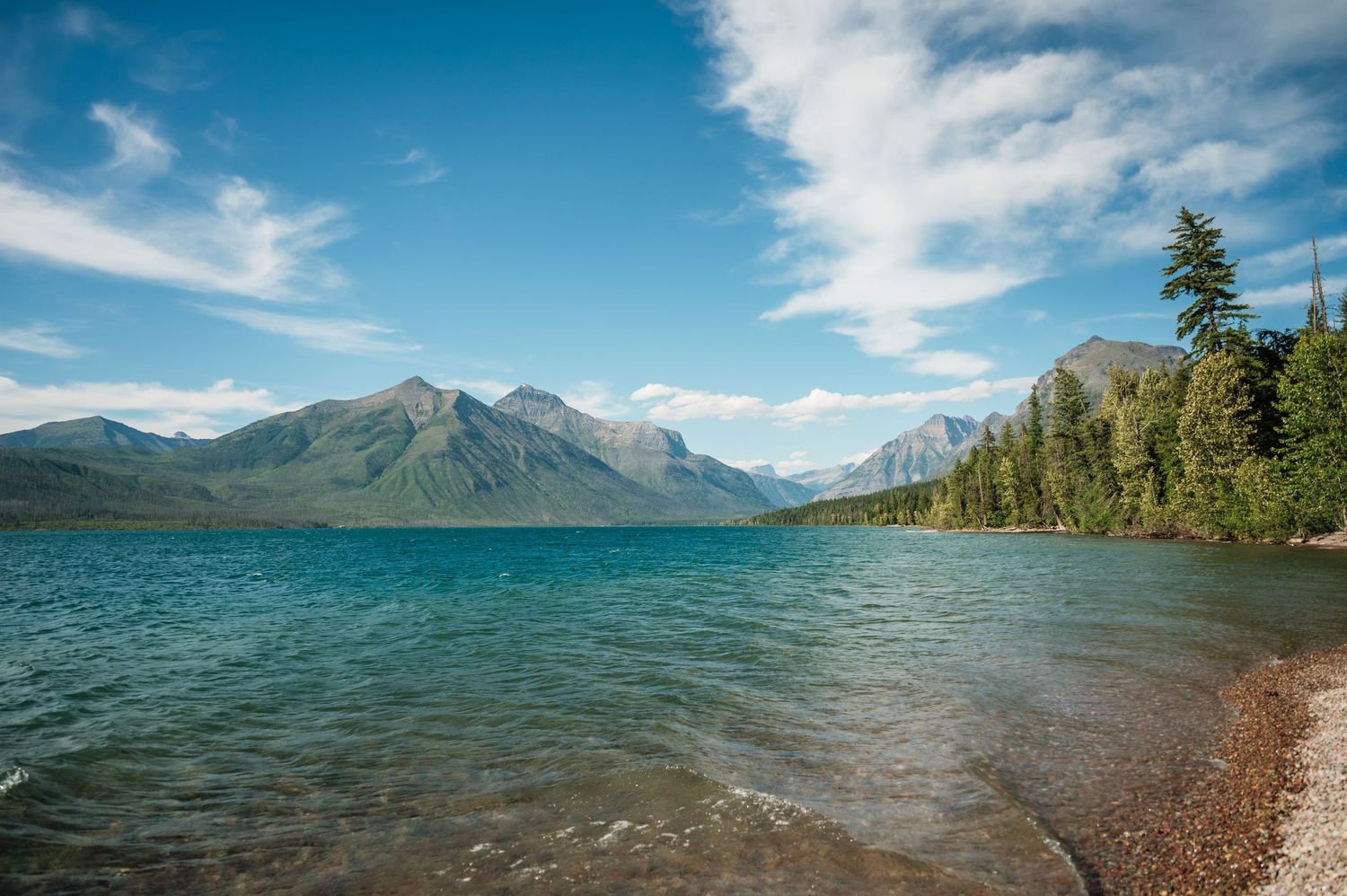 Serene turquoise lake with forested shoreline and snow-capped mountains under a blue sky in Glacier National Park.