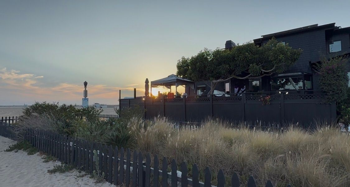 Beachside restaurant with dark fencing at sunset, surrounded by tall grasses and overlooking a serene coastal view.