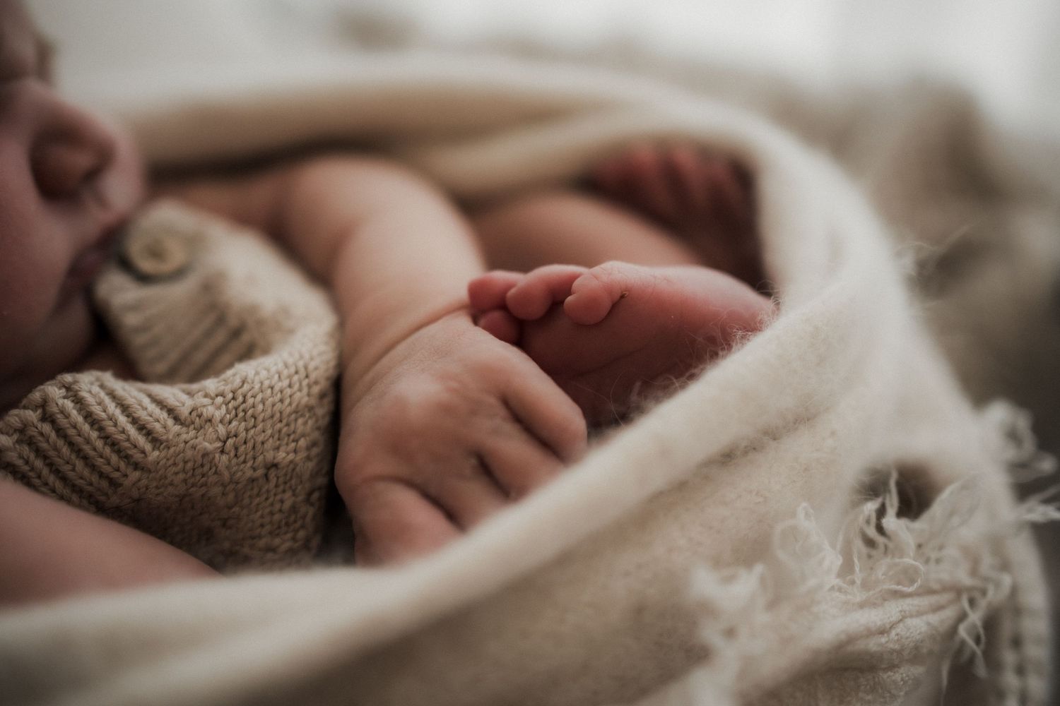 A newborn baby sleeping peacefully wrapped in a soft beige knit blanket, with tiny feet and hands visible.