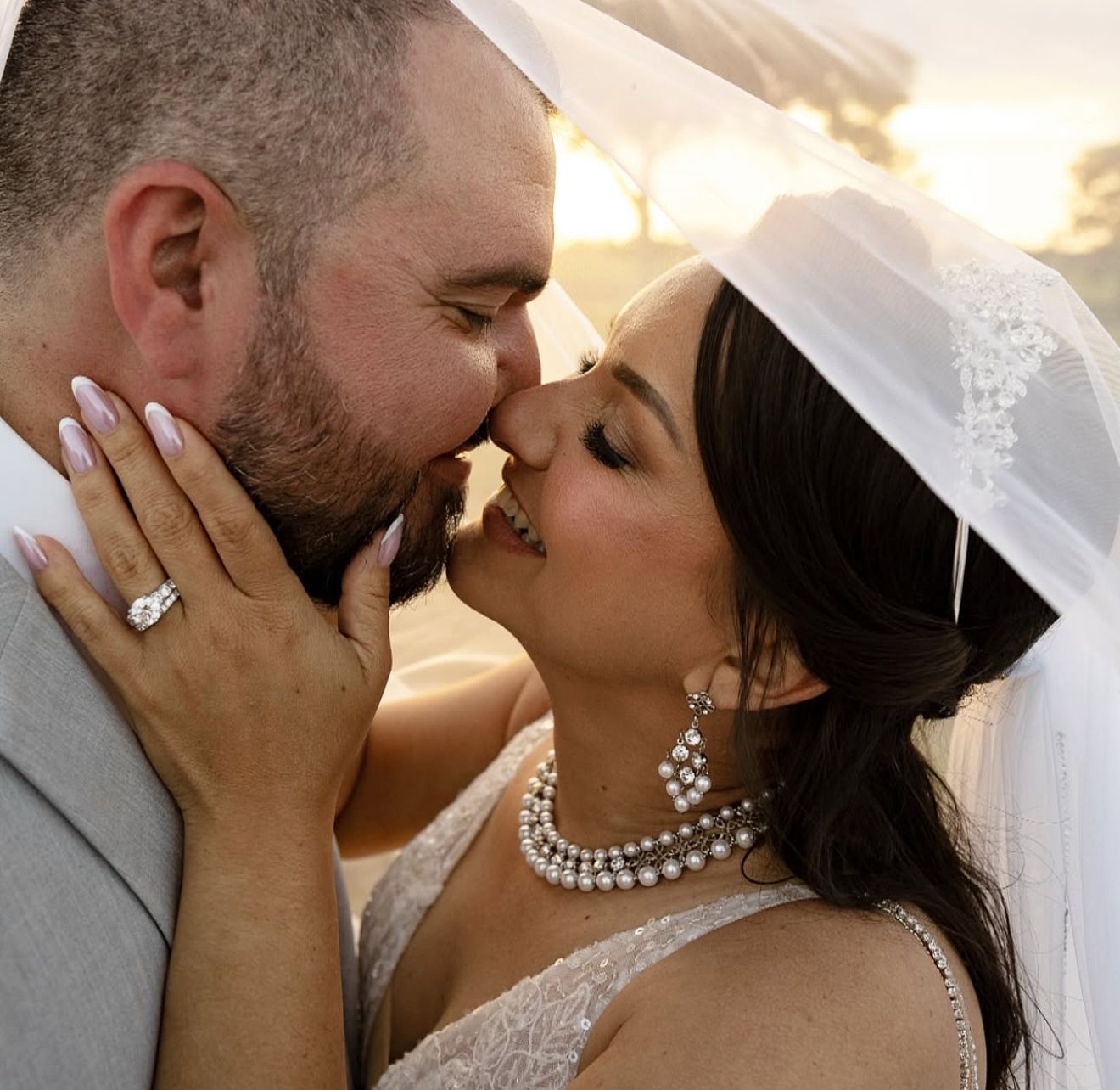 A bride and groom share a romantic kiss at sunset, bride wearing a veil and pearl necklace on their wedding day.