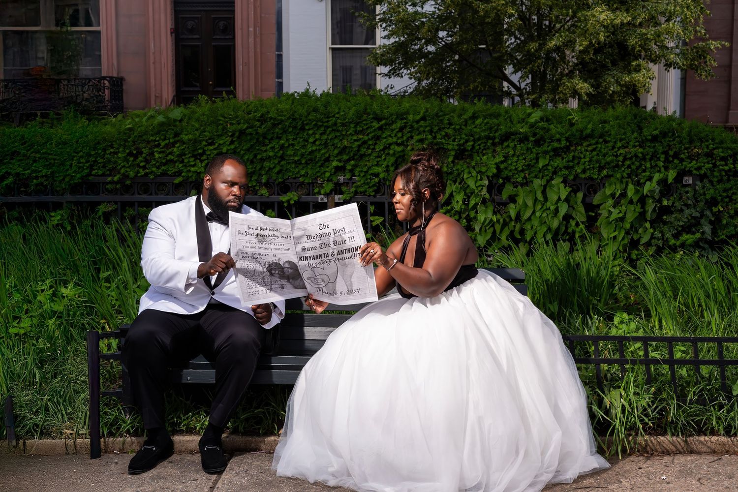A couple in wedding attire sits on a bench reading a newspaper together in front of lush green hedges.