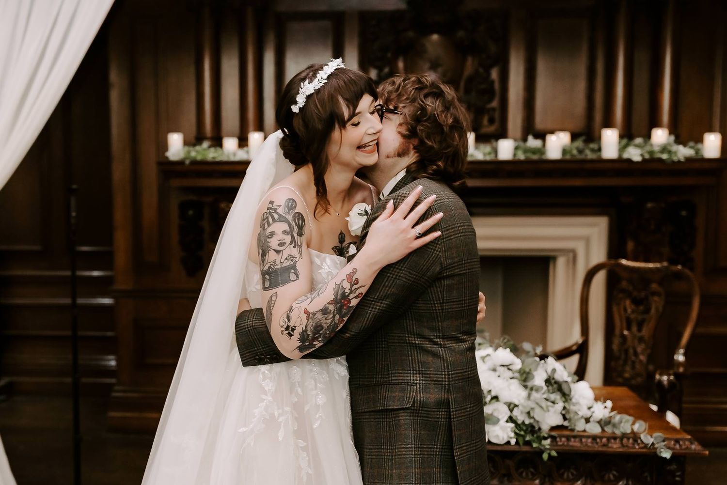 A joyful couple embraces and laughs during their wedding ceremony in front of a fireplace with candles and floral decor.