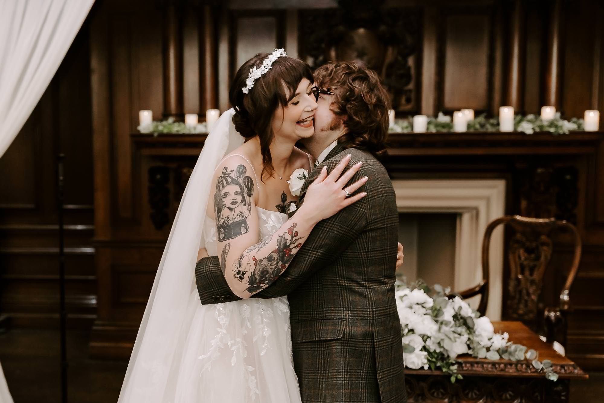 A joyful couple embraces and laughs during their wedding ceremony in front of a fireplace with candles and floral decor.