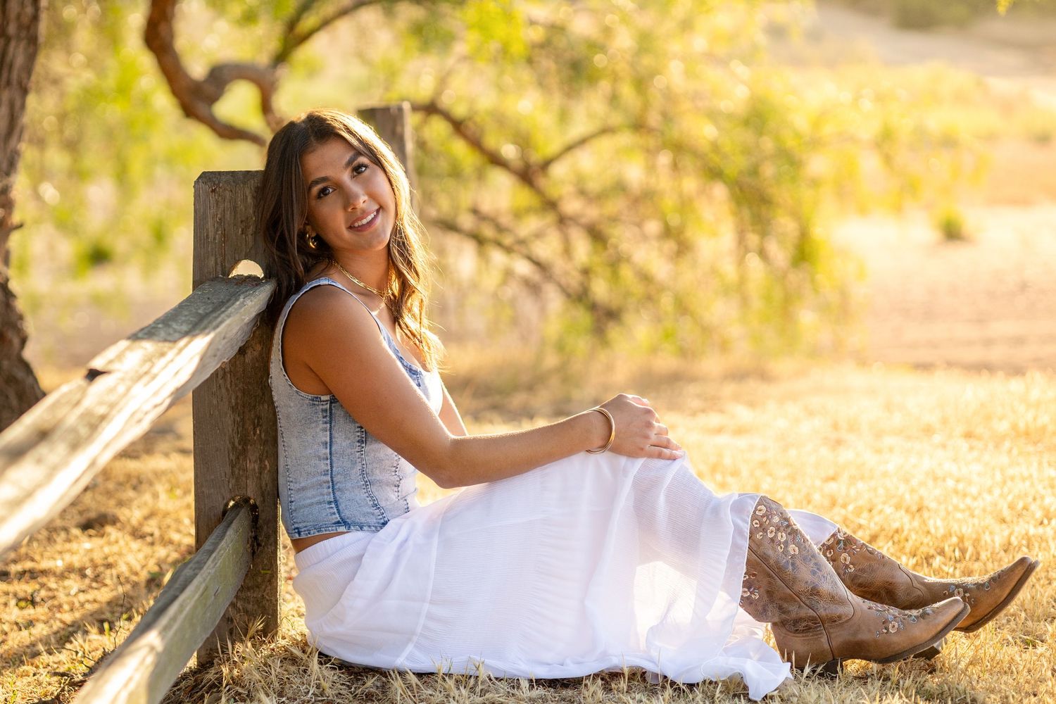 Young woman in white dress and cowboy boots smiling while sitting against a wooden fence in a sunny field.
