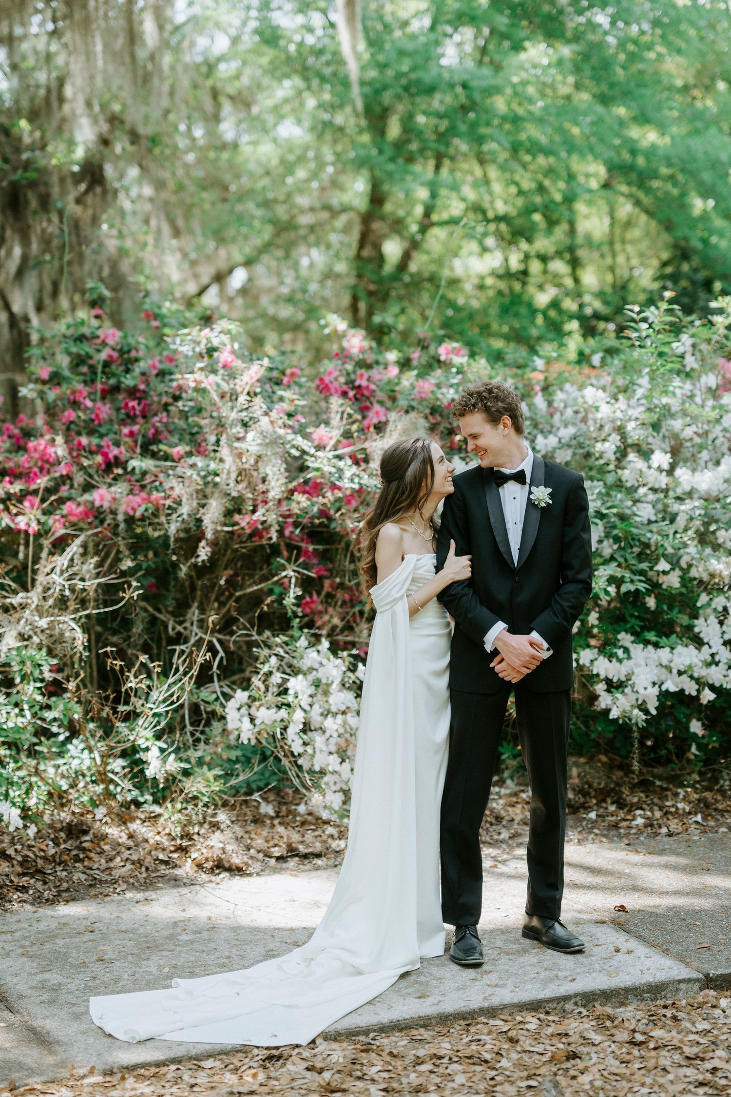 A bride in a white off-shoulder gown and groom in black tuxedo share a romantic moment in a blooming garden.