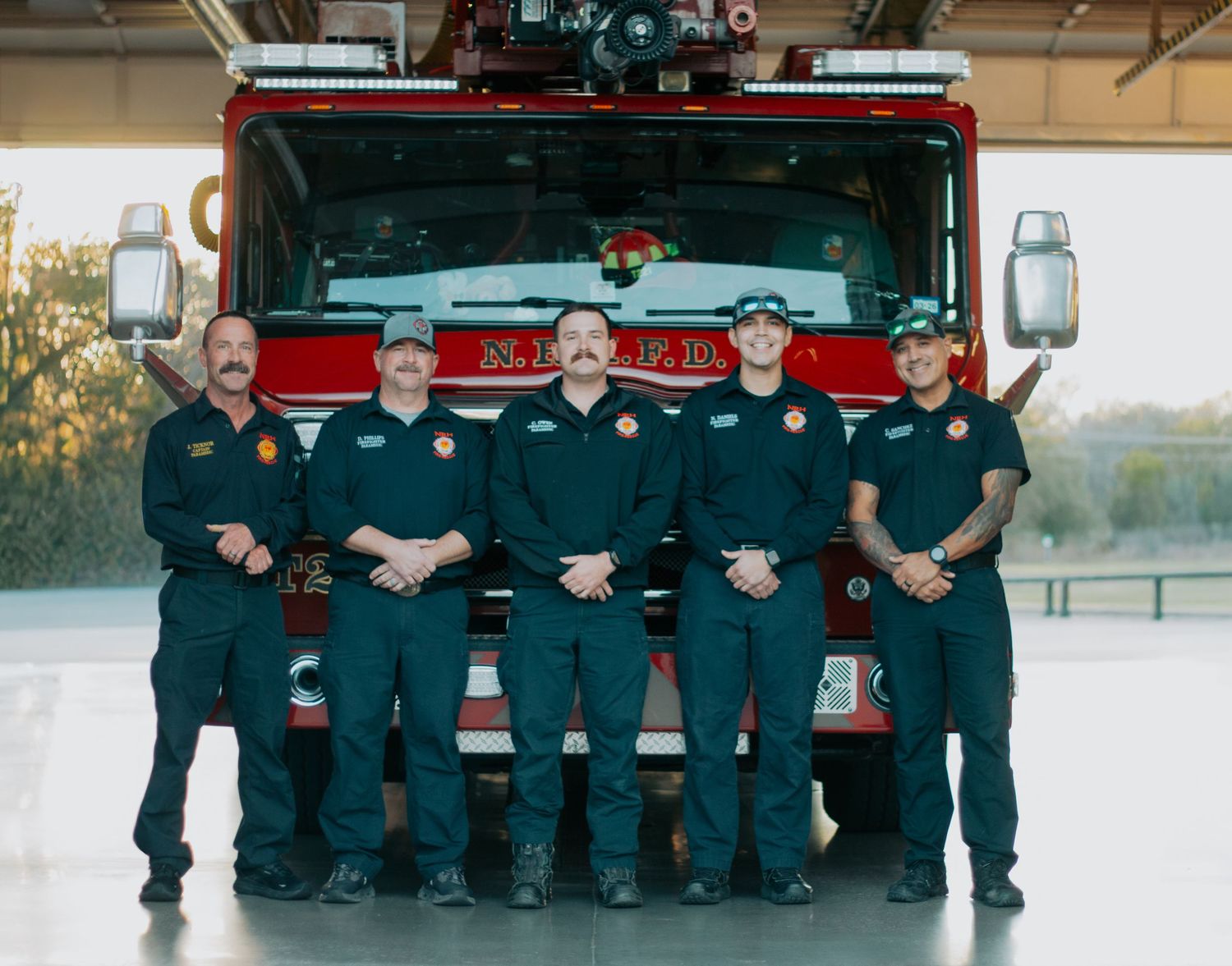 A group of firefighters in navy blue uniforms stand in front of their red fire truck at the station.
