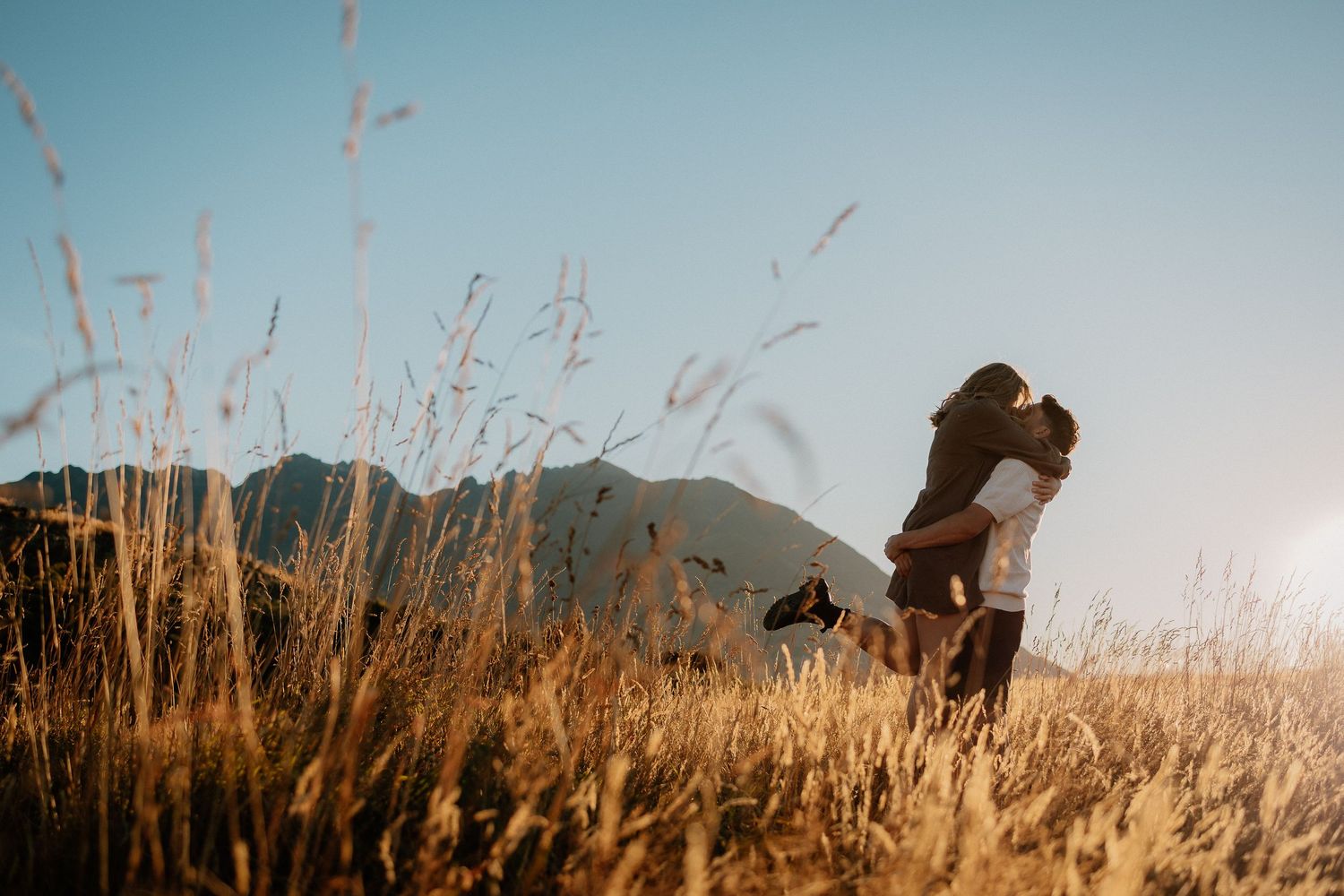 Silhouette of couple embracing in golden wheat field at sunset with mountains in background.