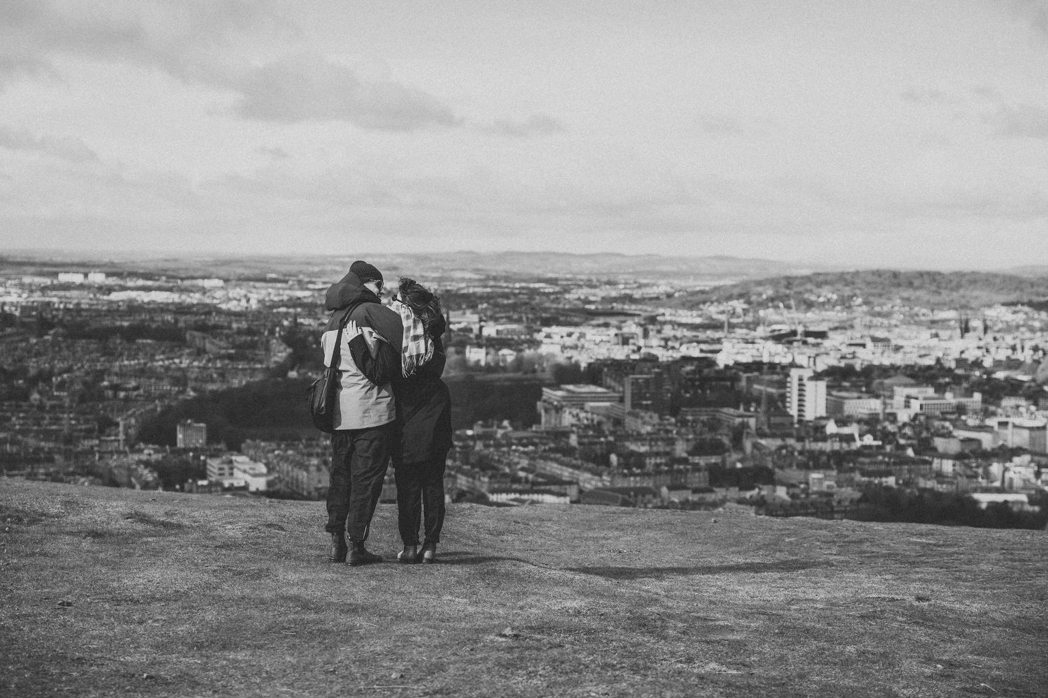 Black and white photo of a couple embracing on a hilltop overlooking a vast cityscape on a cloudy day.