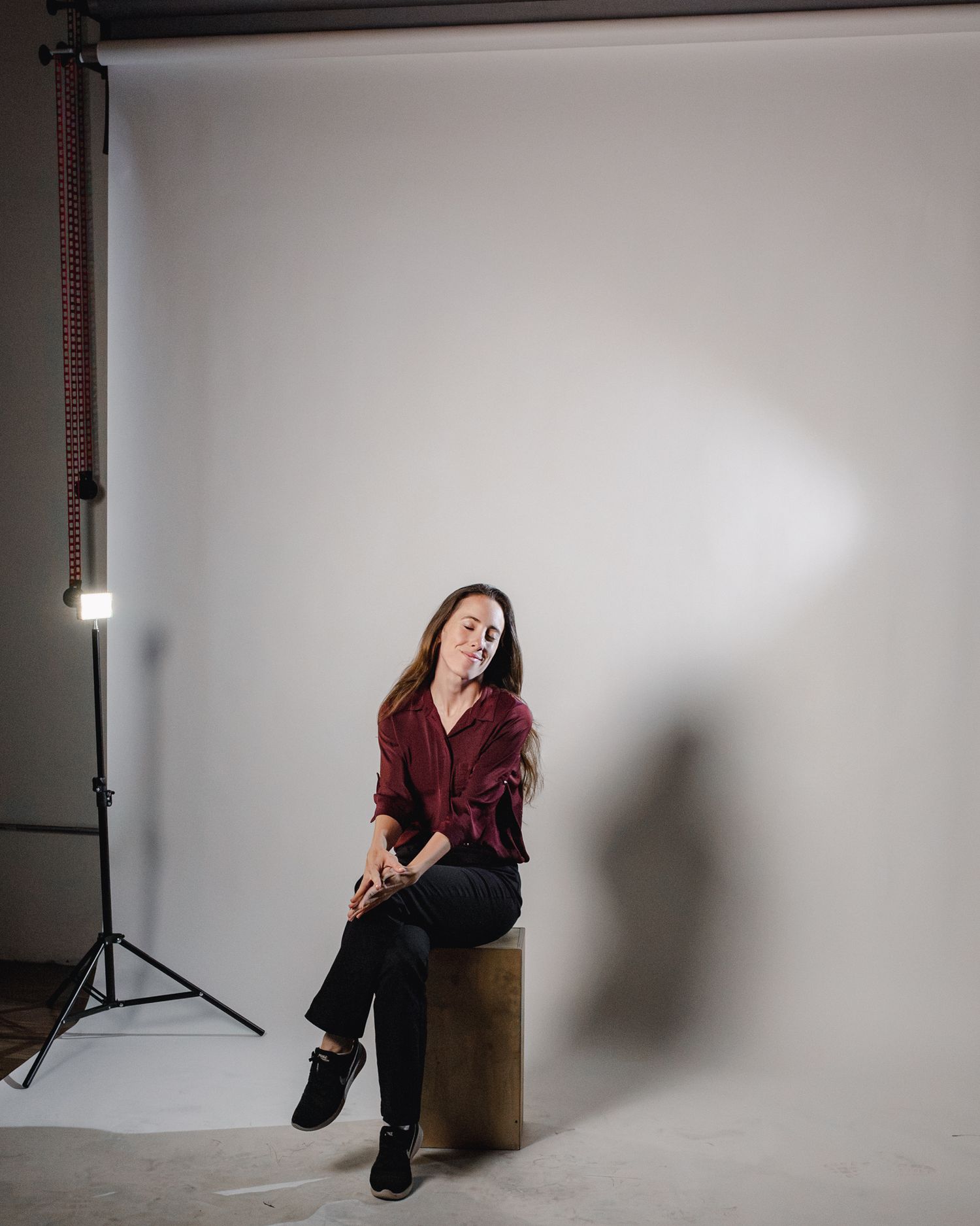 Woman in burgundy shirt and black pants sits on a stool in a photography studio with white backdrop.