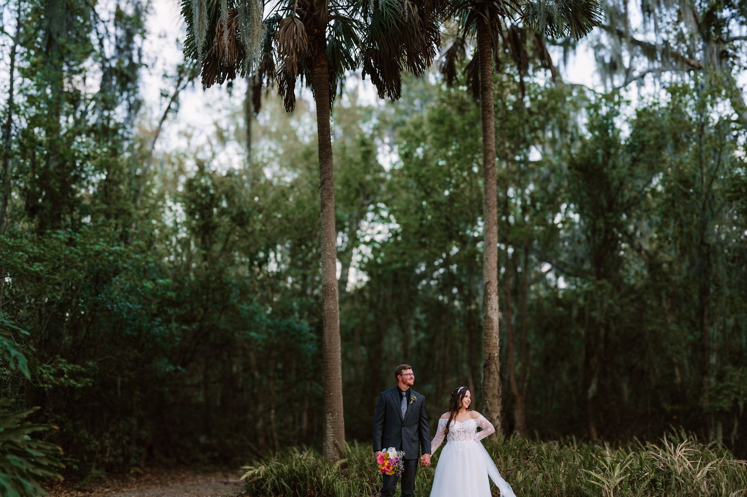 A couple stands beneath Spanish moss hanging from tall trees in a romantic forest setting.