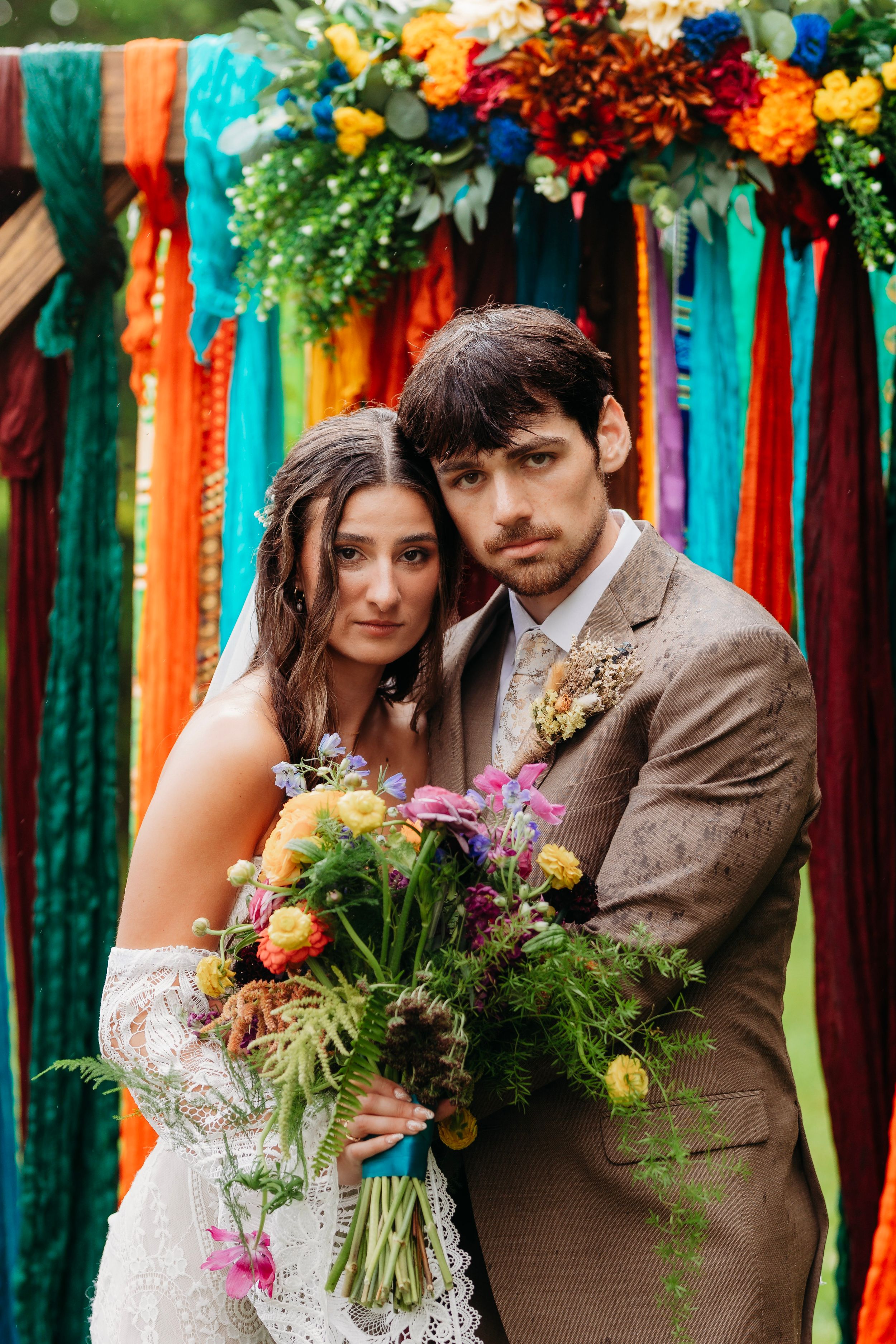 Bride and groom pose before colorful ribbon backdrop with wildflower bouquet at boho outdoor wedding ceremony.