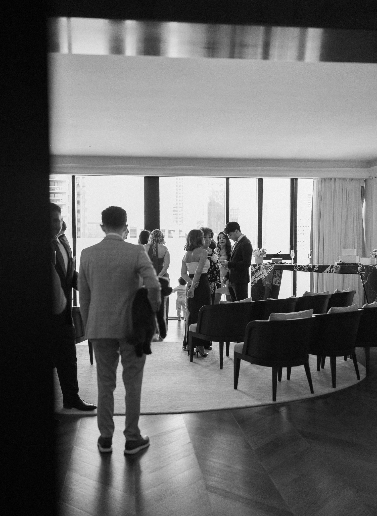 Black and white photo of elegantly dressed guests mingling in a modern high-rise event space with city views.