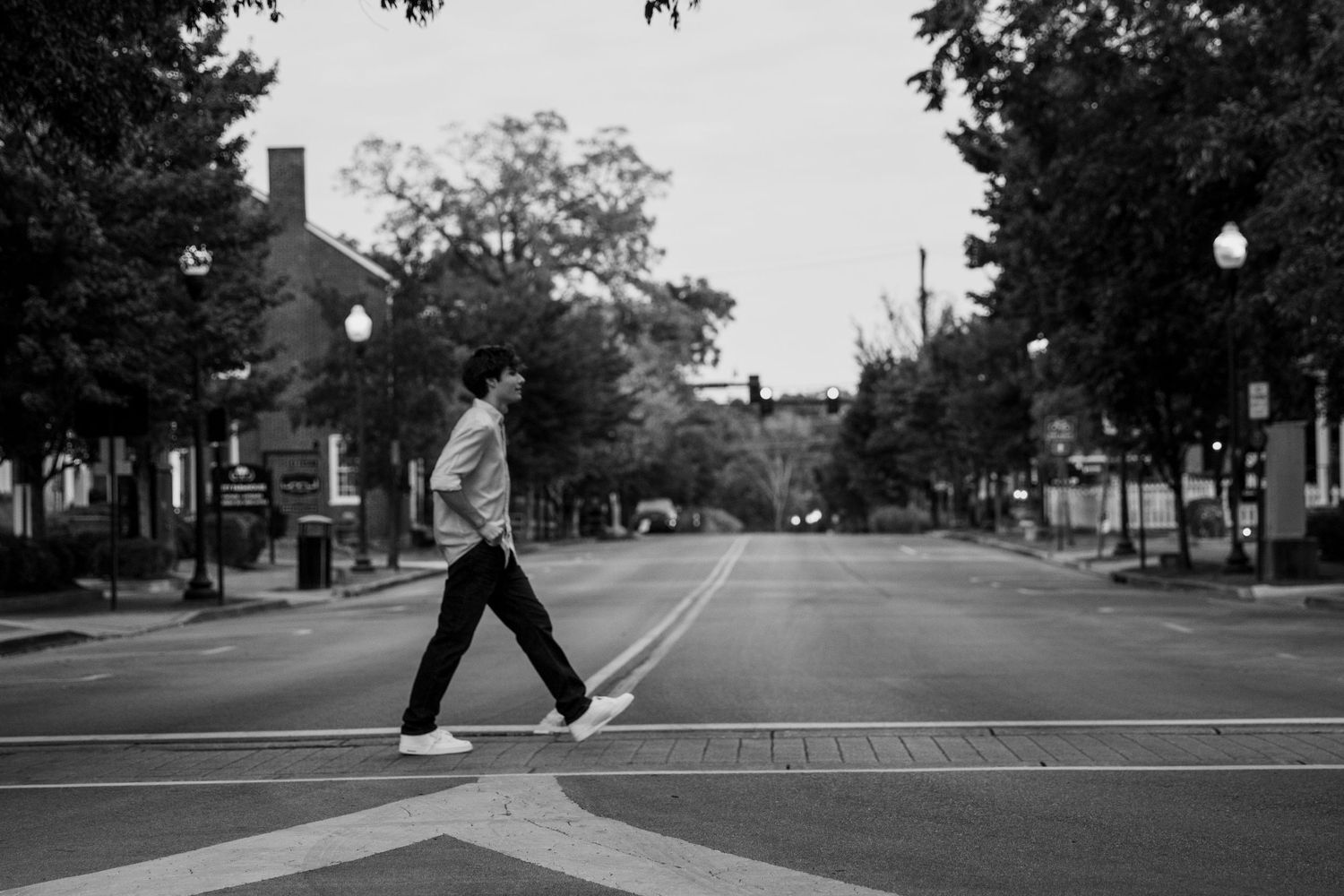 Black and white photo of a person crossing an empty urban street lined with trees and historic buildings.