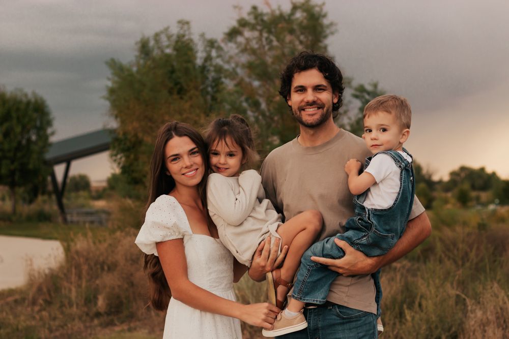 Happy young family of four posing outdoors near a pond with lush green trees and a dramatic cloudy |Westminster Colorado Family Photographer