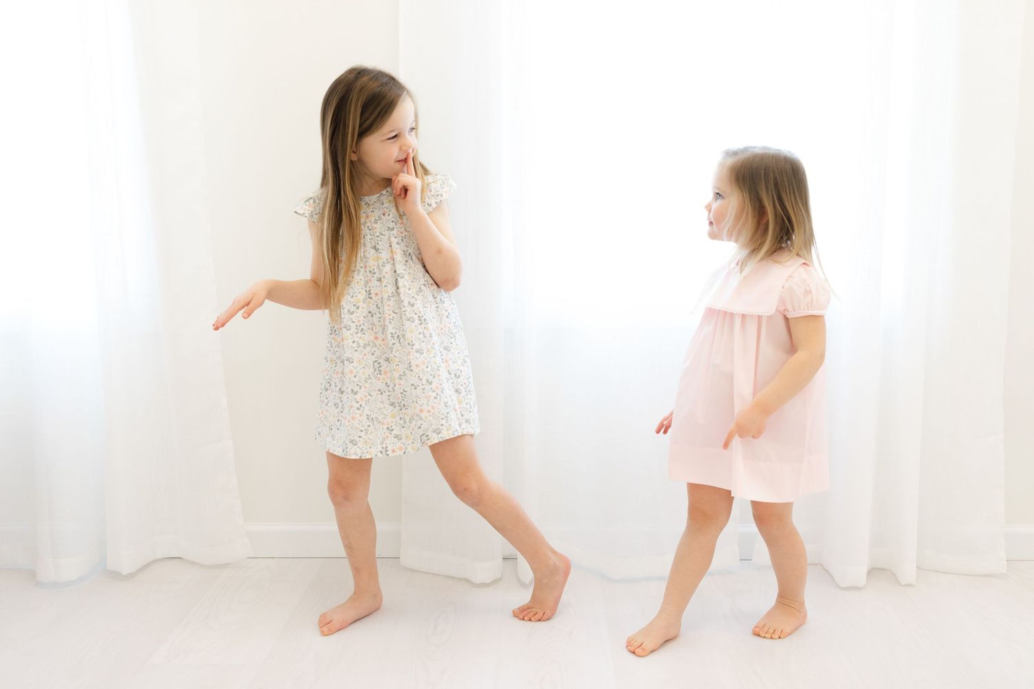 Two young girls in dresses playing and dancing together in a bright white room. Easy ways to document family pictures in Peachtree City, GA.