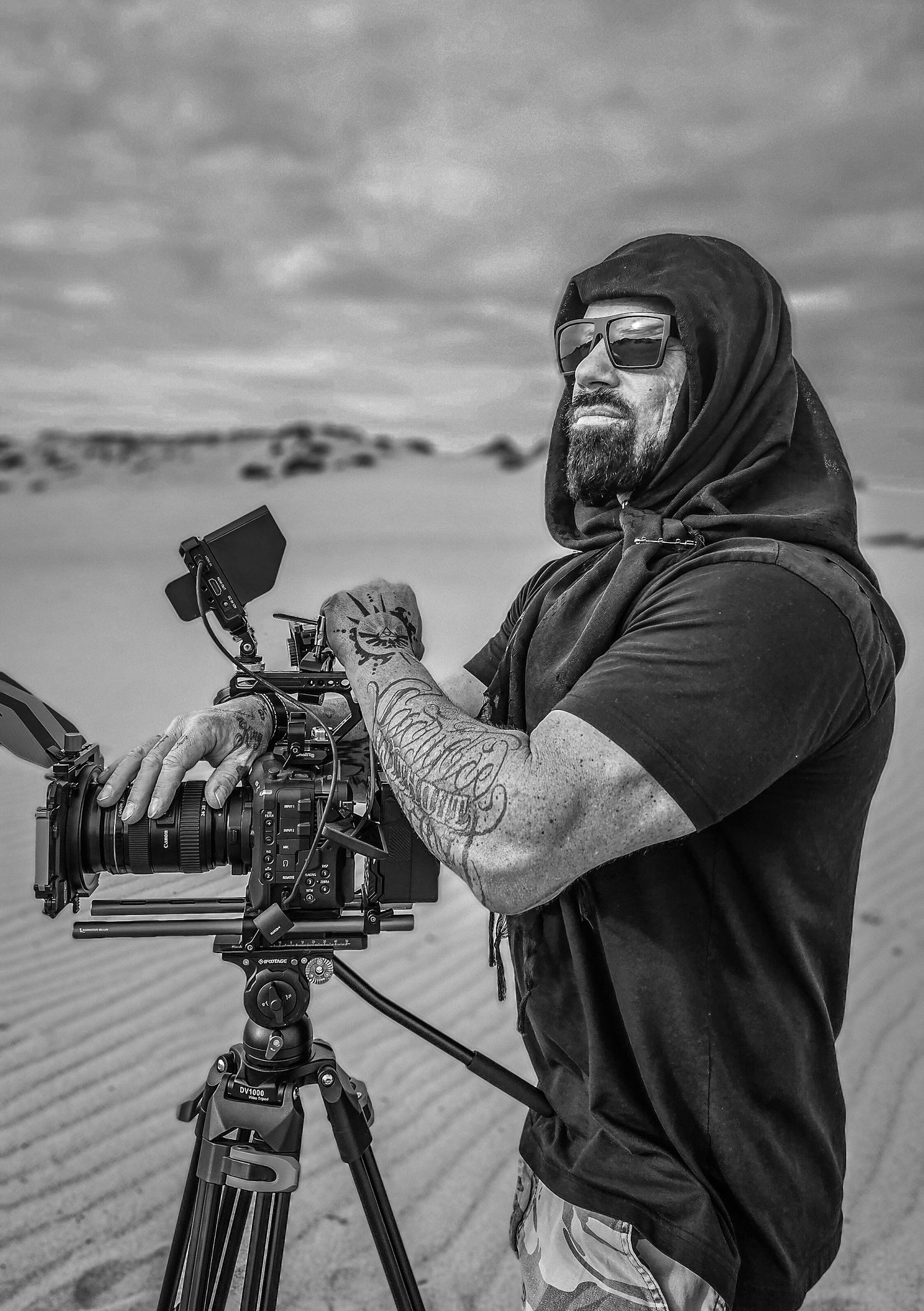 Professional videographer in black hoodie operates camera on tripod in dramatic black and white composition.