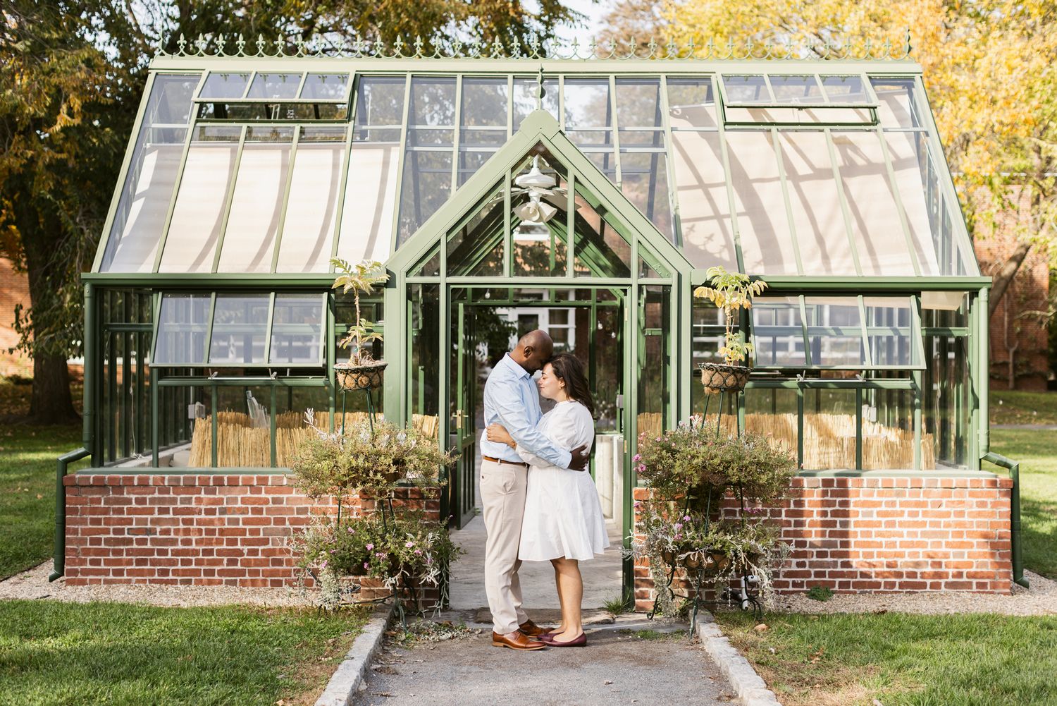 Couple embraces each other in front of a glass greenhouse at Garden at Elm Bank in Wellesley, Massachusetts for couple photos.