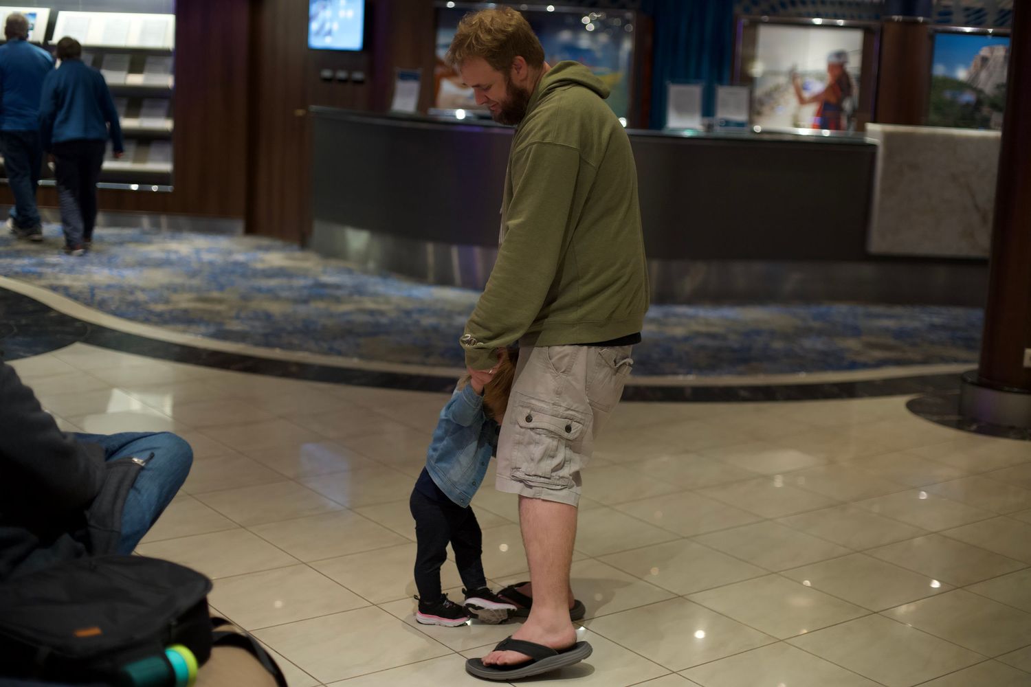 Man in green hoodie and flip flops bending down to interact with a small child in a hotel lobby.
