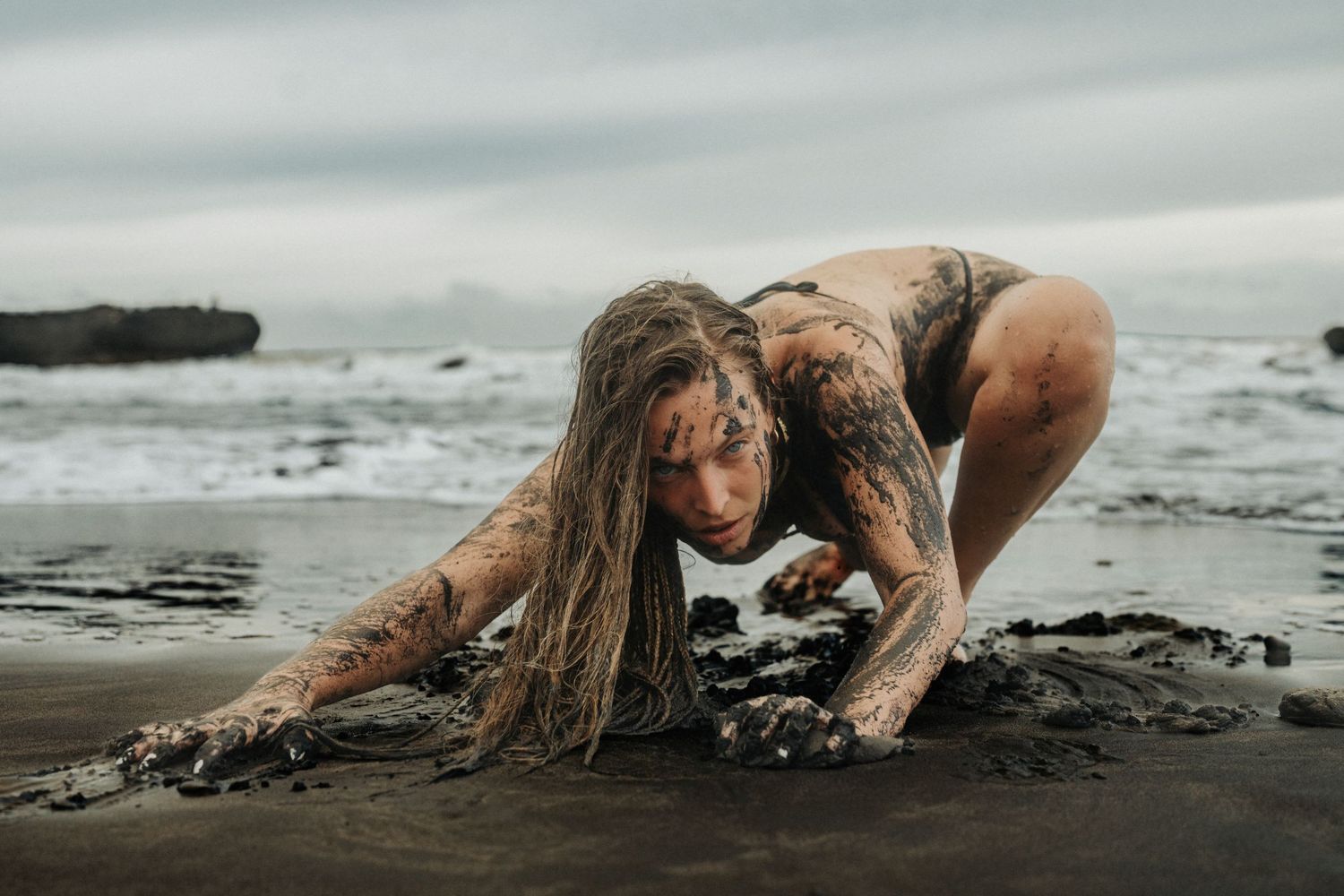 Woman covered in dark mud crouching on a black sand beach with dramatic ocean waves in the background.