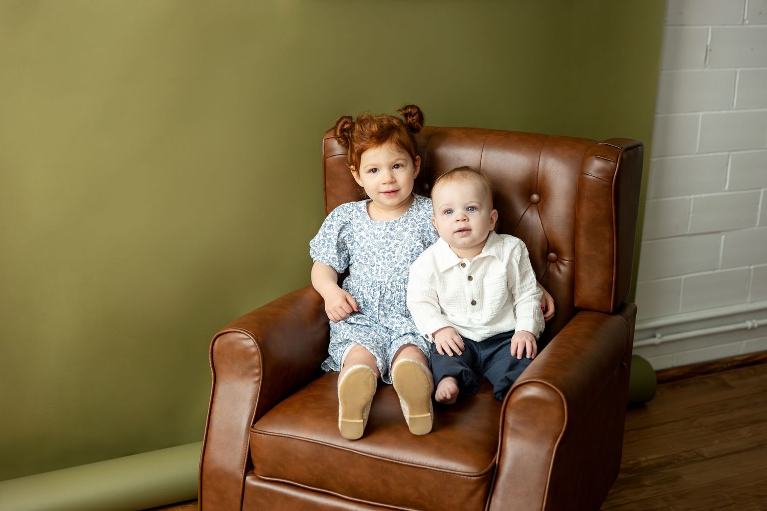 Two young siblings sit together in a brown leather tufted armchair against an olive wall.