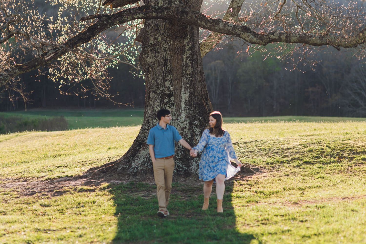 Couple holding hands under a large tree in a sunny green park, woman in blue floral dress, man in khaki pants.