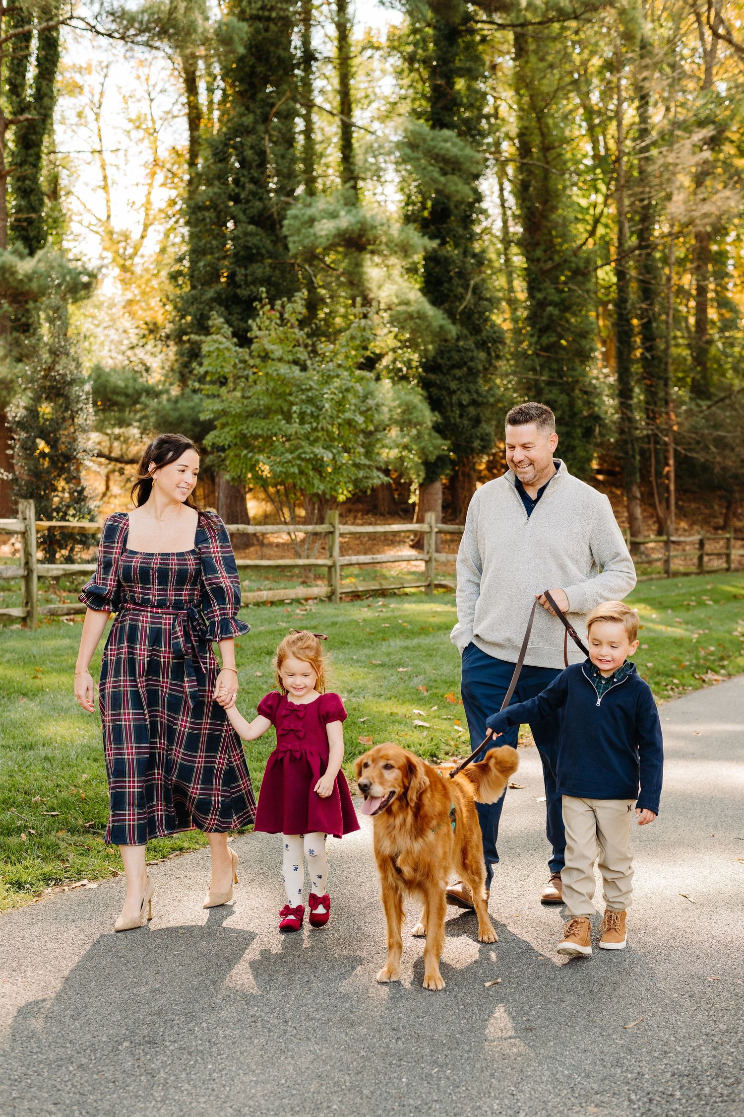 A family walks together on a paved path in a wooded park with their golden retriever dog on an autumn day.